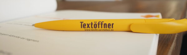 Im Fokus ist ein gelber Kugelschreiber abgebildet. Mit dem lilanem Schriftzug Textöffner.