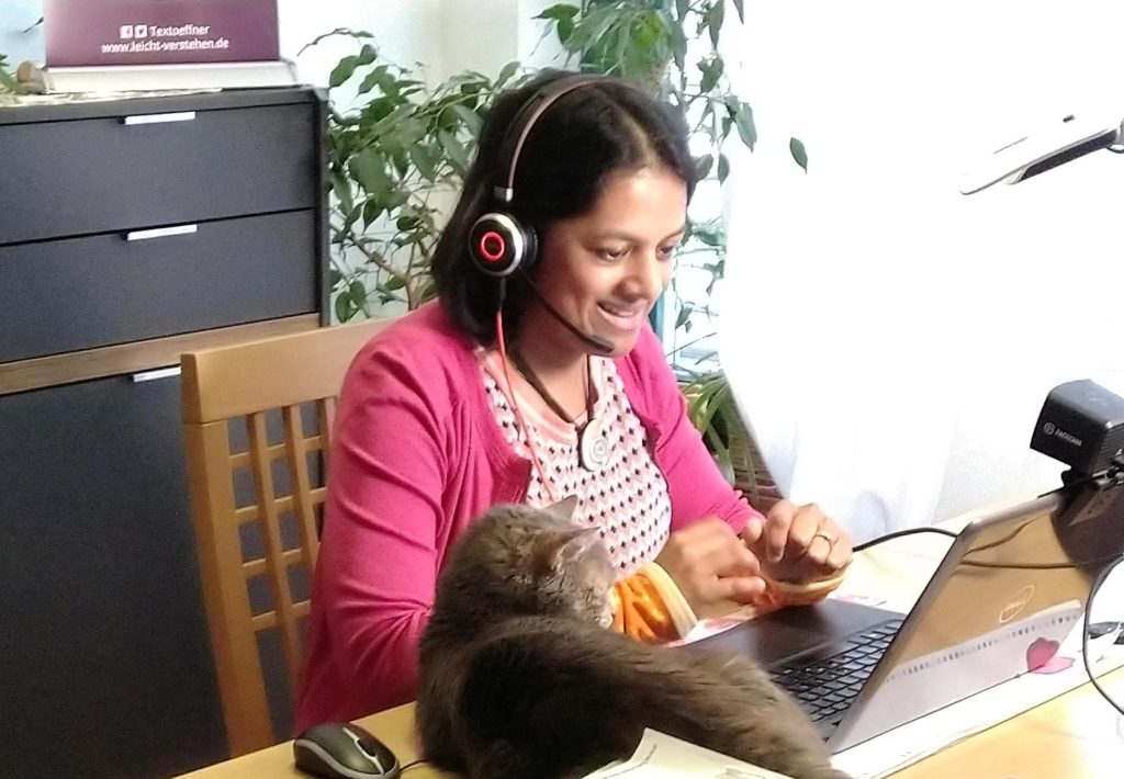Krishna-Sara Helmle sitzt auf einem Stuhl. Vor ihr auf dem Tisch steht ihr Laptop. Ihre Blickrichtung ist auf den Bildschirm gerichtet. Auf ihrem Kopf trägt sie ein Headset. Auf dem Bildschirm ist eine Webcam befestigt. Krishna-Sara Helmle lächelt in die Kamera. Neben dem Laptop liegt ihre Katze.