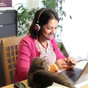 Krishna-Sara Helmle sitzt auf einem Stuhl. Vor ihr auf dem Tisch steht ihr Laptop. Ihre Blickrichtung ist auf den Bildschirm gerichtet. Auf ihrem Kopf trägt sie ein Headset. Auf dem Bildschirm ist eine Webcam befestigt. Krishna-Sara Helmle lächelt in die Kamera. Neben dem Laptop liegt ihre Katze.