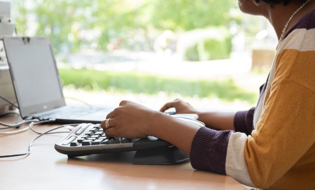 Krishna-Sara Helmle sitzt an ihrem Schreibtisch und tippt mit ihrer Tastatur einen Text. Sie trägt einen gelb, weiß, lila, gestreiften Pullover.