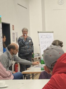 Eine Frau mit weißen kurzen Haaren steht neben einem Flipchart. Sie hält ein Mikrofon in der Hand, das sie an den Mund hält und spricht vor dem Publikum. Auf dem Flipchart steht "Netzwerk Leichte Sprache". 