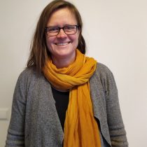 Porträt AK Das Bild zeigt eine Frau. Die Frau hat braune, lange Haare und blaue Augen. Sie hat eine Brille. Sie trägt einen gelben Schaal und eine graue Woll-Jacke.